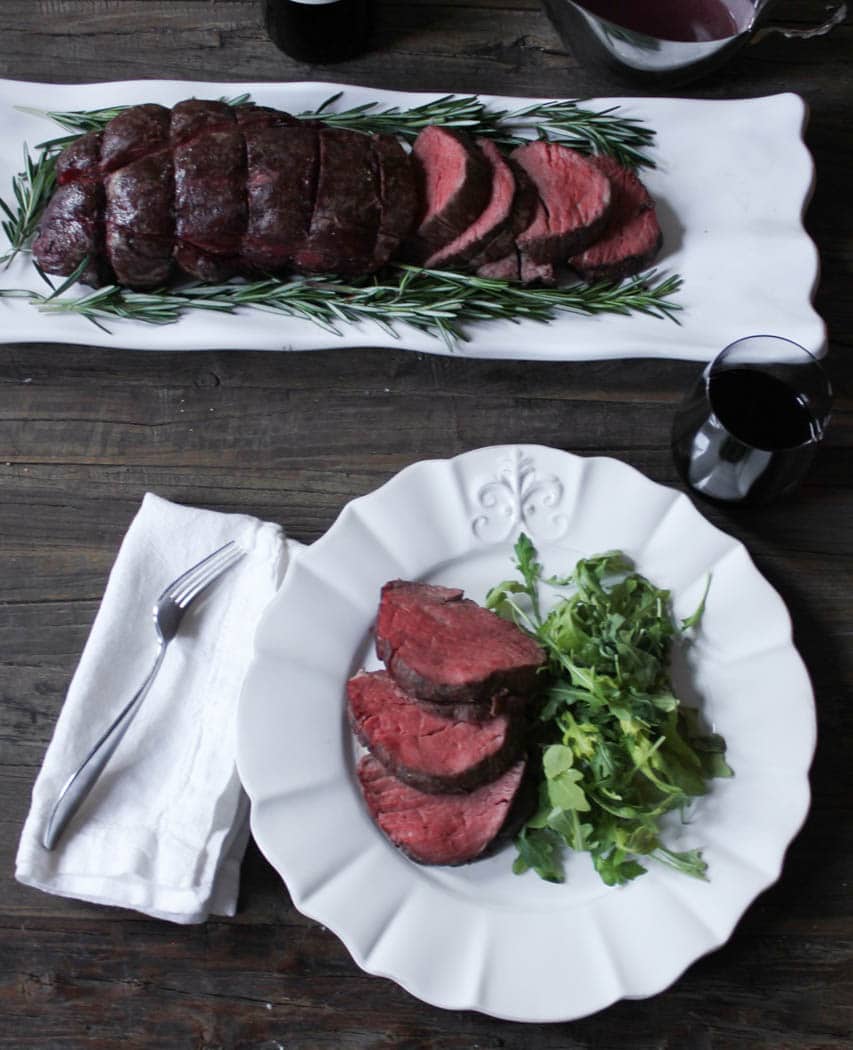 Overhead view of sliced beef tenderloin with rosemary on a plater and one plated serving. 