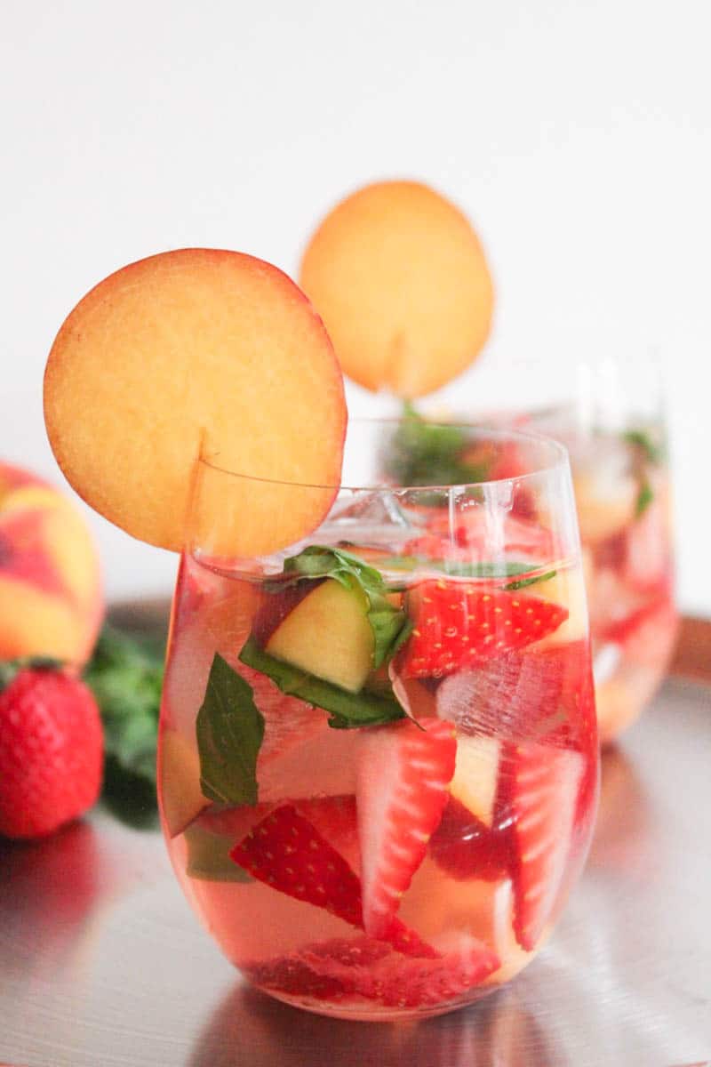 Close-up sideview of one glass of sparkling rose sangria garnished with a peach round with a second glass in the background.