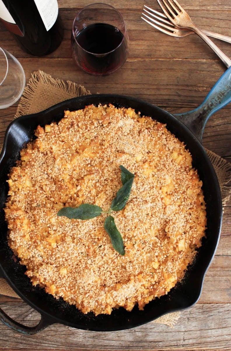 Overhead view of a brown rice bake with sausage and apples in a skillet with two glasses of red wine in the background.