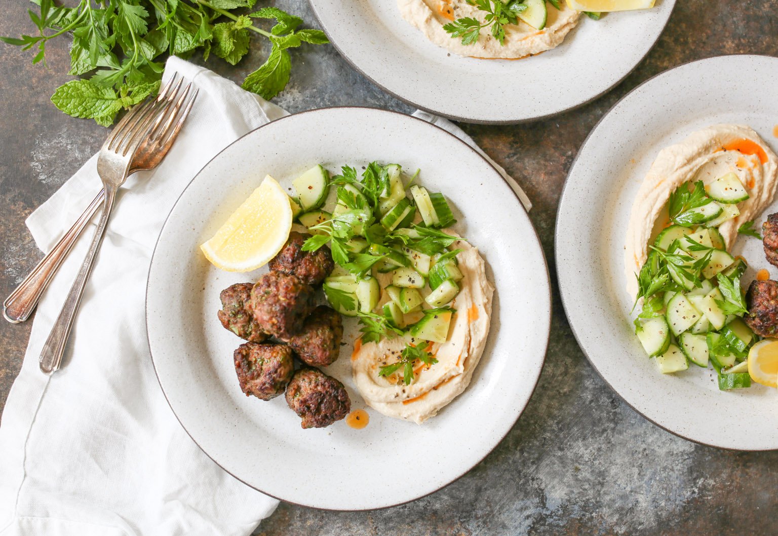 Overhead view of plated lamb kofta with hummus, cucumber salad, and lemon with napkins and forks.