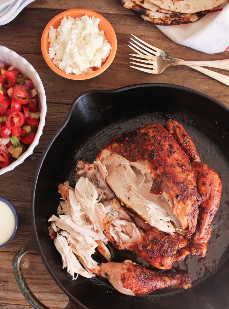Overhead view of a whole slow-roasted chicken with some meat pulled from the bone surrounded by Mediterranean taco fixings. 