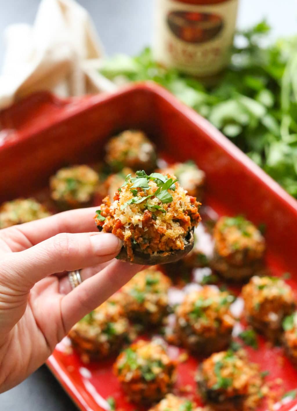 Close up of a hand holding a single stuffed mushroom with a full tray of stuffed mushrooms blurred in the background. 