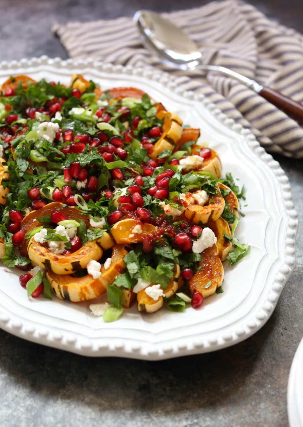 Side view of roasted delicata squash topped with herbs, goat cheese, and pomegranate on a white serving platter with a serving spoon and napkin in the background.